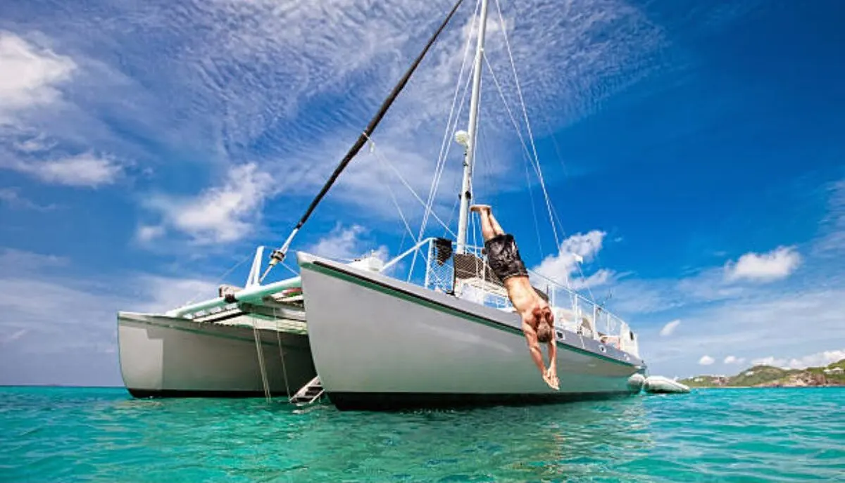 Side view angle of a Catamaran boat with a muscular man diving into the clear sea.