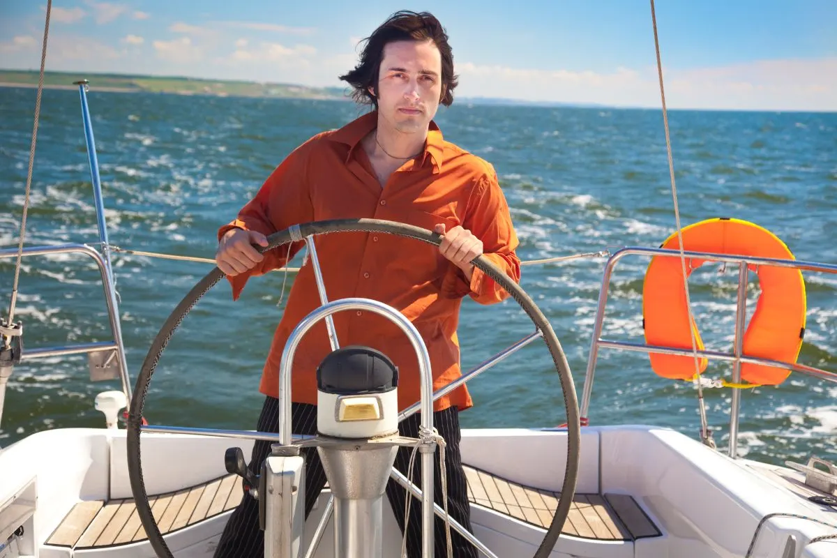 A simple young man is a sailboat captain wearing orange longsleeves navigating his sailboat through the steering wheel.