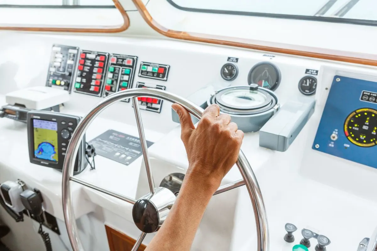 A hand of a man holding his boat's steering wheel in the boat's navigation controls cockpit.