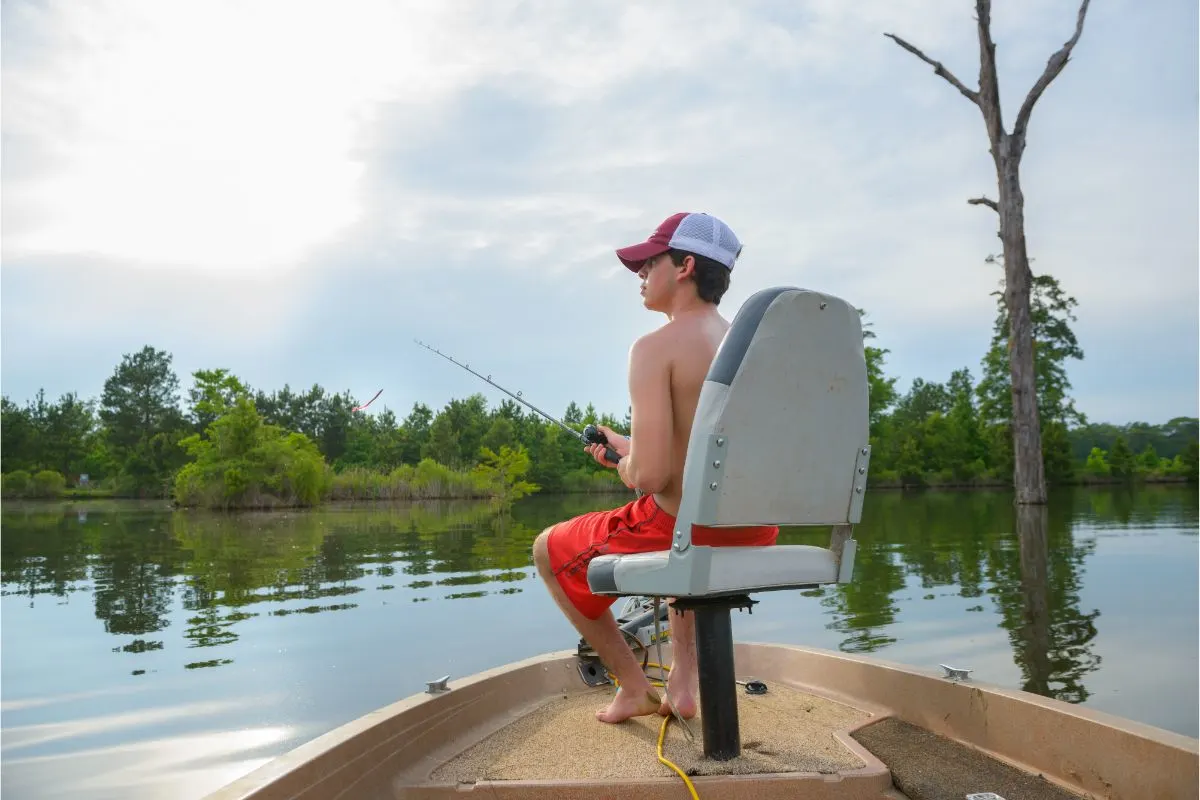 A boy on the boat sitting on a casting seat fishing in the lake.